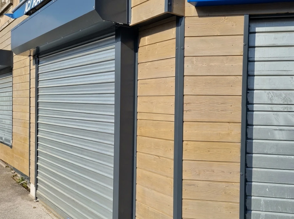 Façade commerciale extérieure combinant bardage bois horizontal en sapin naturel et volets roulants métalliques gris anthracite